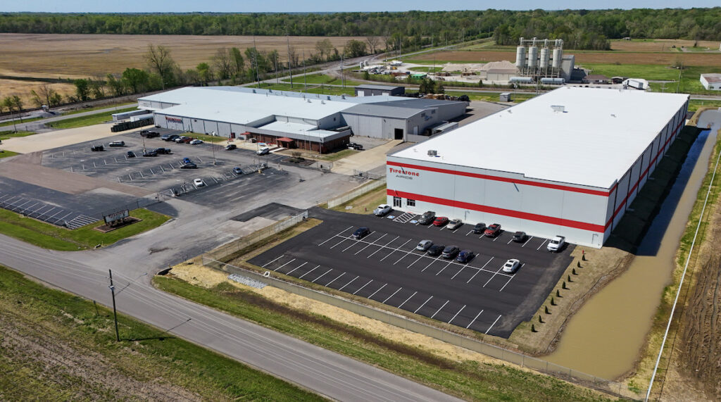 Aerial view of an industrial complex with large warehouse buildings, parking lots with several cars, and surrounding grassy fields. The complex is bordered by roads and a small pond is visible near the foreground.