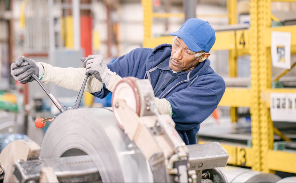 A factory worker in safety glasses, gloves, and a blue cap operates heavy machinery, adjusting a large roll of metal in an industrial setting with shelves and equipment in the background.