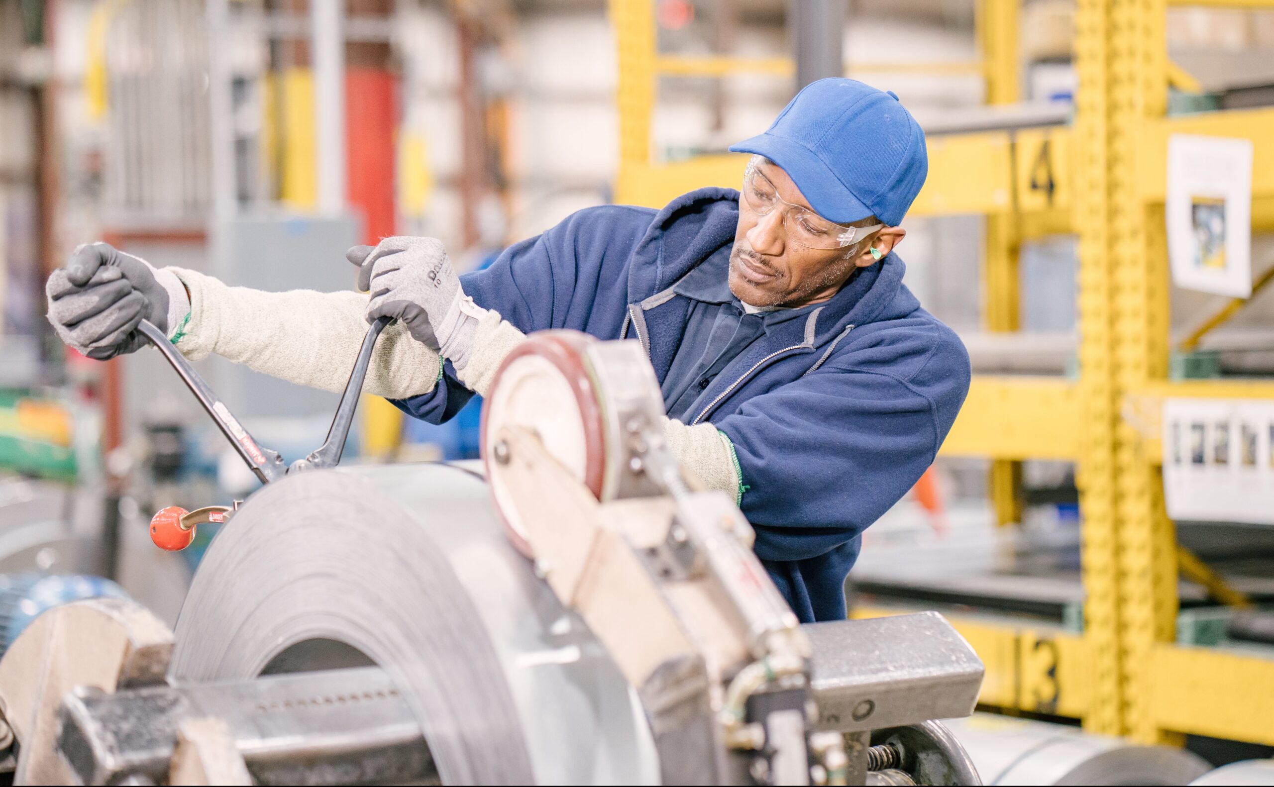 A factory worker in safety glasses, gloves, and a blue cap operates heavy machinery, adjusting a large roll of metal in an industrial setting with shelves and equipment in the background.