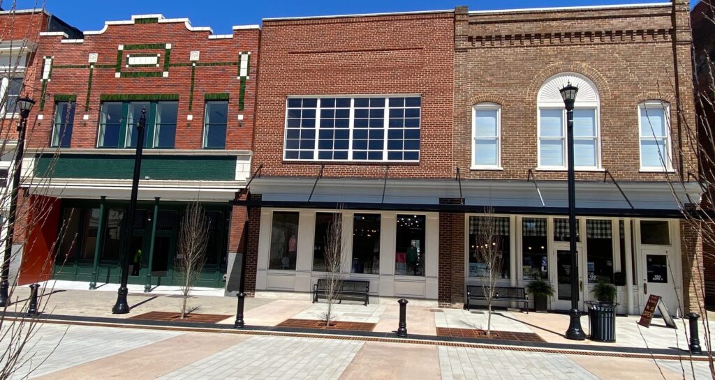 Two brick storefronts on a sunny day, featuring large windows and awnings. The sidewalk in front is wide with young trees and black street lamps, and no people are visible.