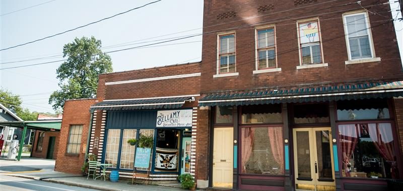 A small-town street features brick buildings with large windows; one has a sign reading "Bellamy Cafe" and colorful chairs outside, while the other storefront has closed doors and curtains. Power lines cross above.