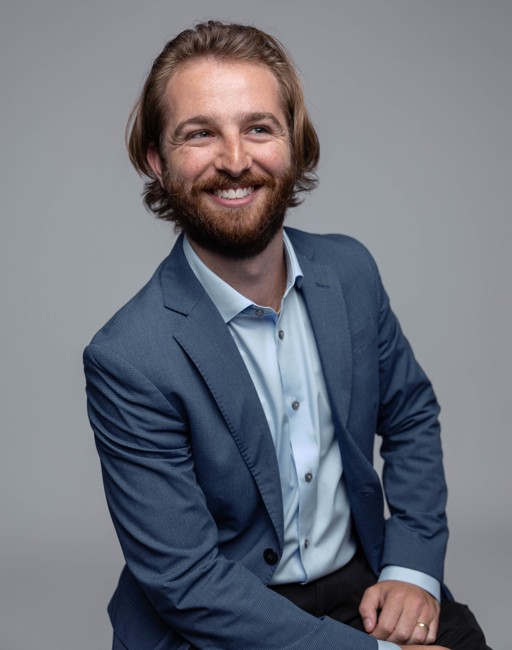 A man with light brown hair and a beard, wearing a blue suit and light blue shirt, smiles while sitting against a plain grey background.