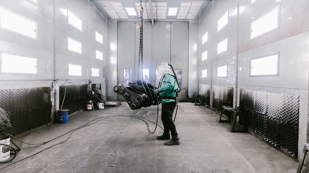 A person wearing protective gear stands in an industrial room, operating a suspended heavy machine part with a chain hoist. The space has metallic walls, bright ceiling lights, and industrial equipment.