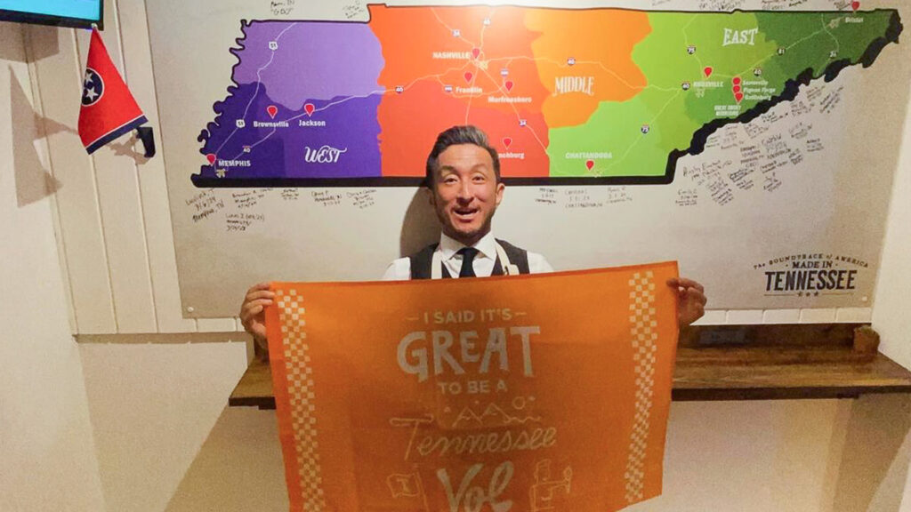 A man stands smiling indoors, holding an orange sign that reads "I said it’s great to be a Tennessee Vol," in front of a colorful map of Tennessee on the wall.