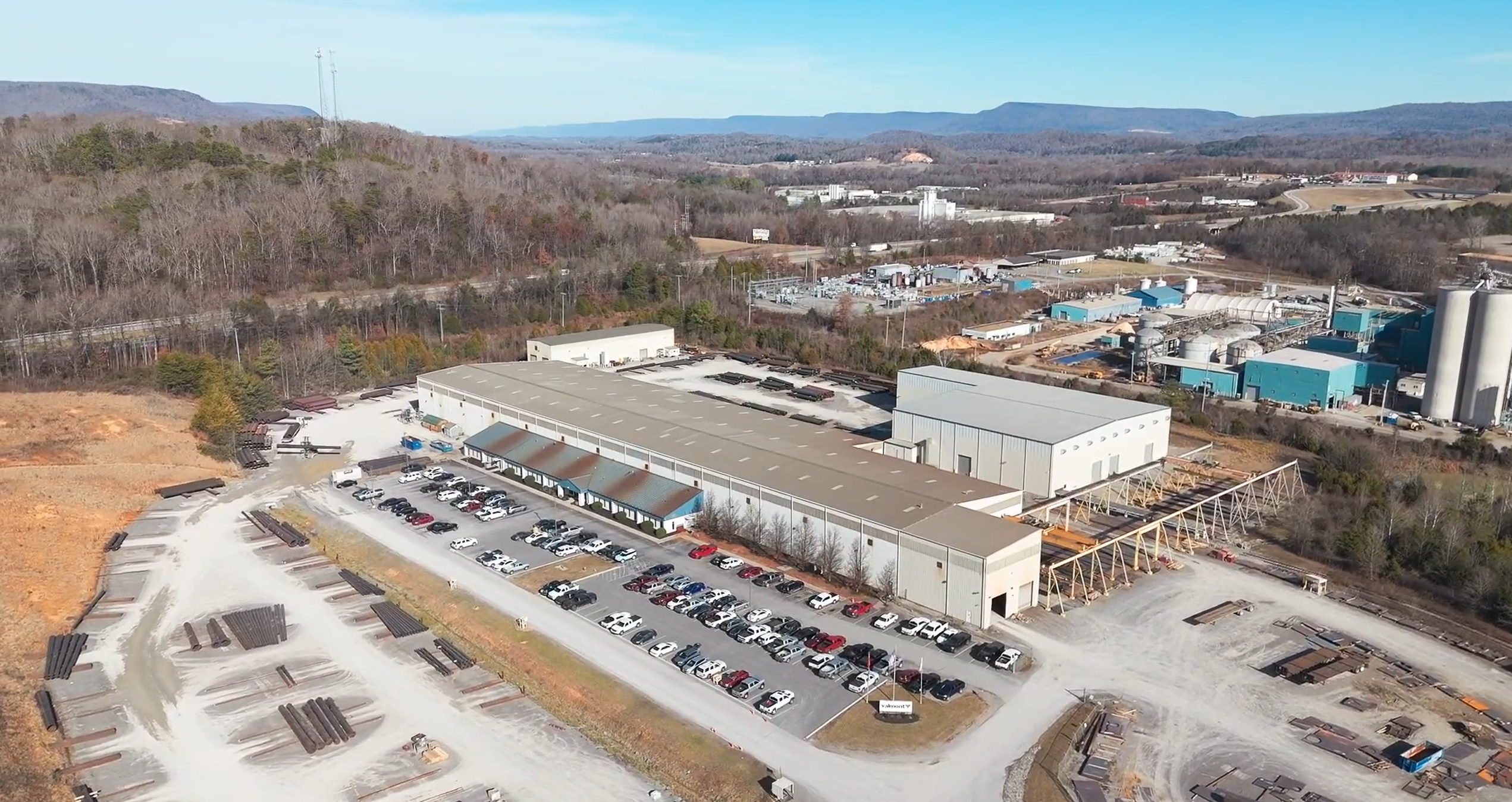 Aerial view of an industrial facility with two large warehouse buildings, a parking lot filled with cars, and surrounding woods and hills under a clear sky. Industrial materials and structures are visible around the site.