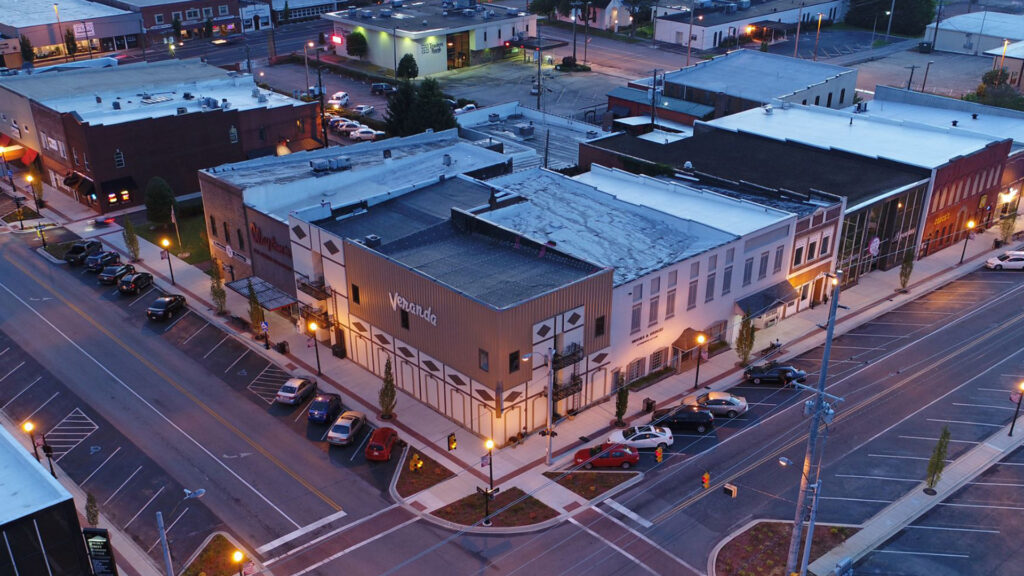 Aerial view of a small town’s downtown area at dusk, showing several parked cars along the streets, illuminated streetlights, and brick buildings with businesses on quiet, intersecting roads.