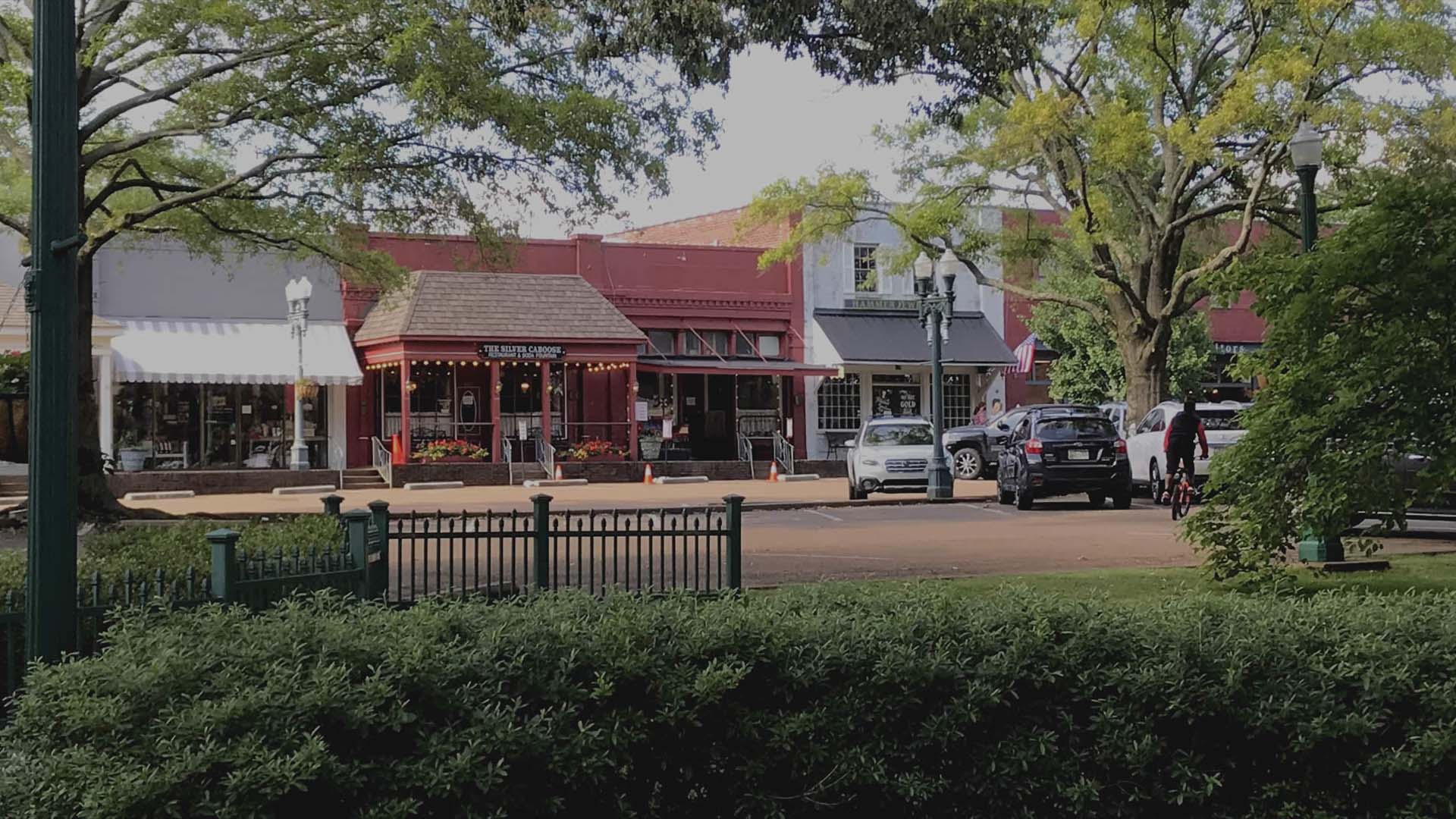 A quaint small-town street with historic storefronts, cars parked along the road, leafy trees providing shade, and a decorative green fence and bushes in the foreground.