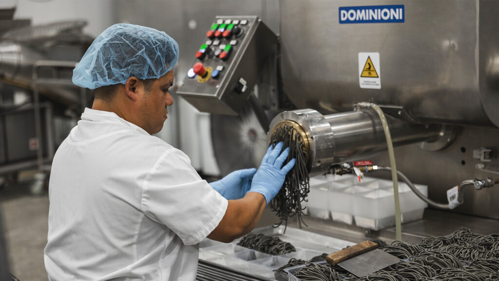 A worker in a white uniform and blue hairnet operates a large industrial pasta machine, guiding dark-colored pasta strands into a tray in a food processing facility.