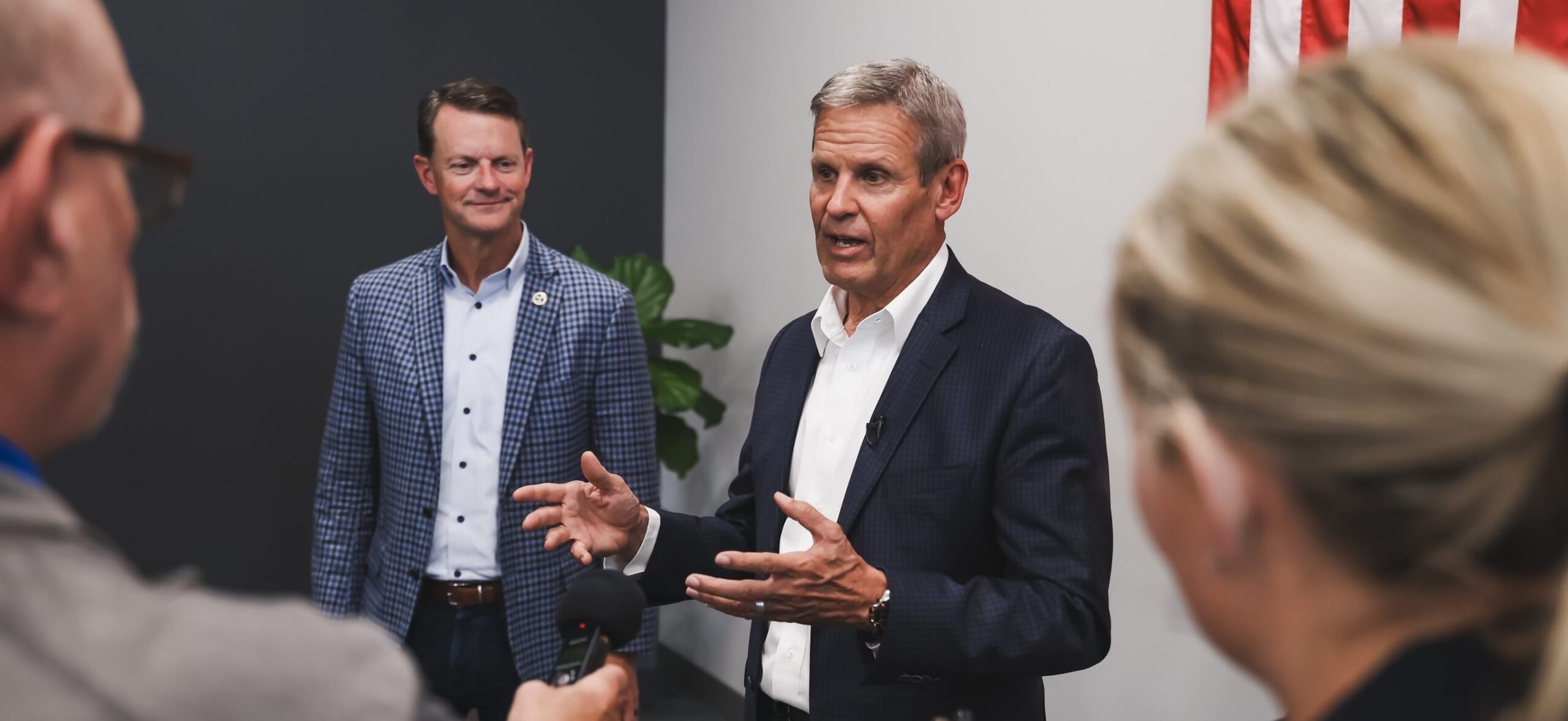 Two men in suits speak to reporters indoors; one man gestures with his hands while the other stands behind him, smiling. A U.S. flag is partially visible on the wall. Reporters hold microphones toward them.