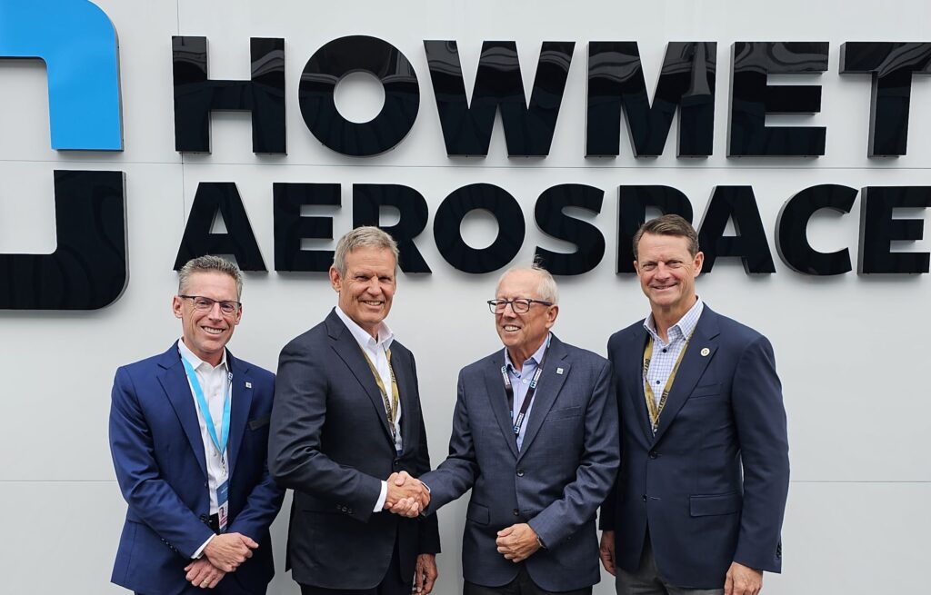 Four men in business attire stand smiling in front of a large Howmet Aerospace sign. Two men in the center are shaking hands, indicating a formal or celebratory occasion.