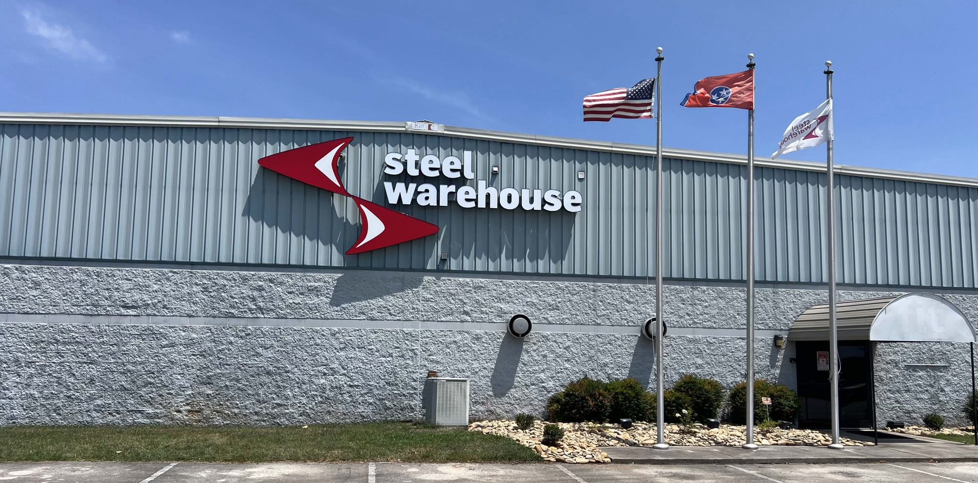 A gray industrial building with a "Steel Warehouse" sign, three flagpoles flying the U.S., Tennessee, and another flag, a small landscaped area, and a blue sky in the background.