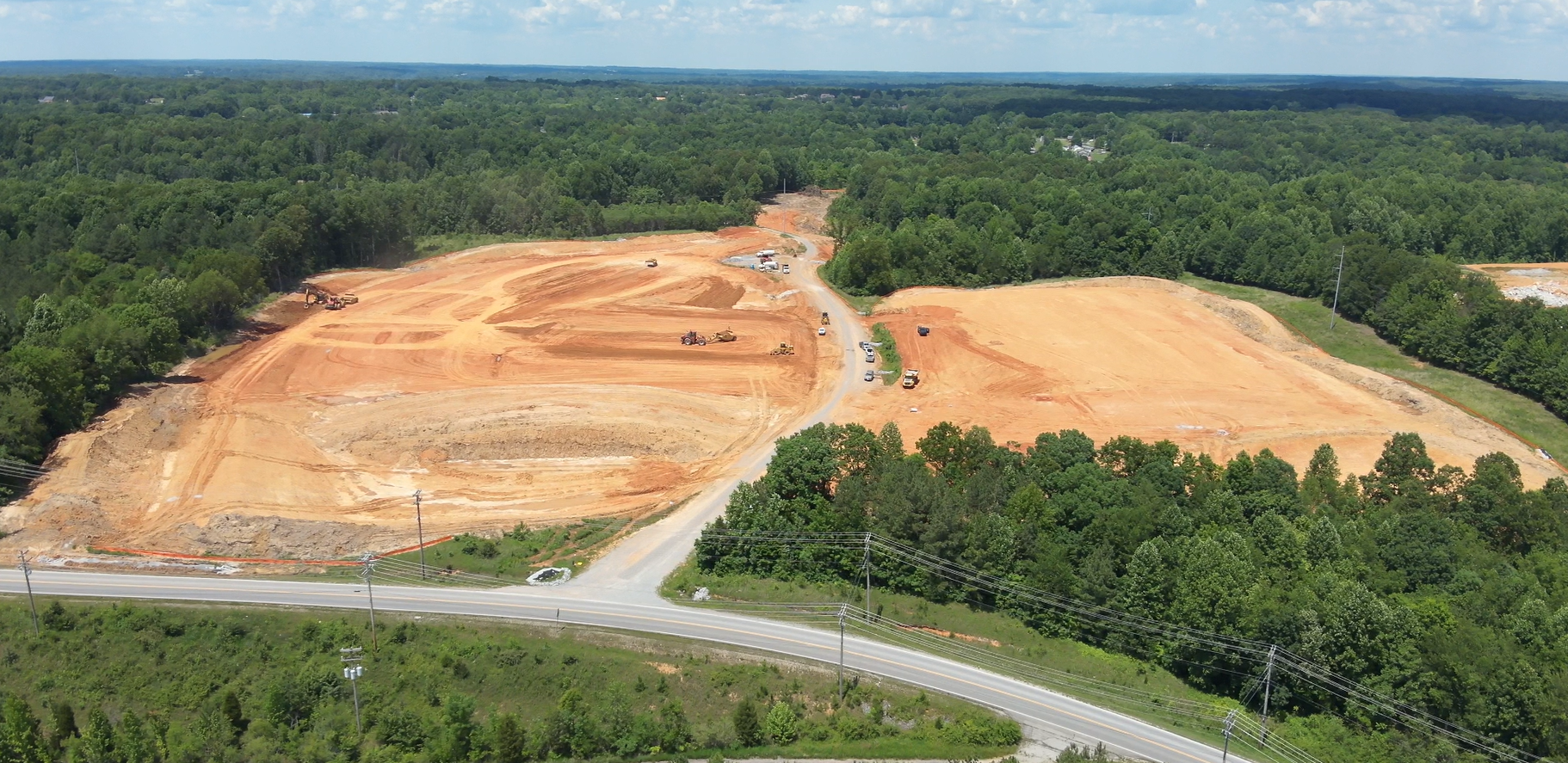 A large construction site with cleared, leveled reddish-brown earth, surrounded by dense green forest. Several vehicles and machinery are visible on the site, with a road and cars in the foreground.