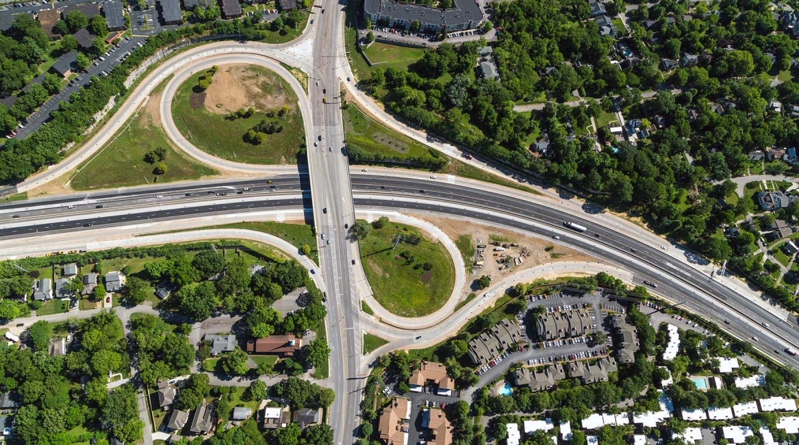Aerial view of a highway interchange surrounded by trees and residential buildings, with curved on-ramps and off-ramps connecting multiple roads. Cars are visible on the roads and parking lots nearby.