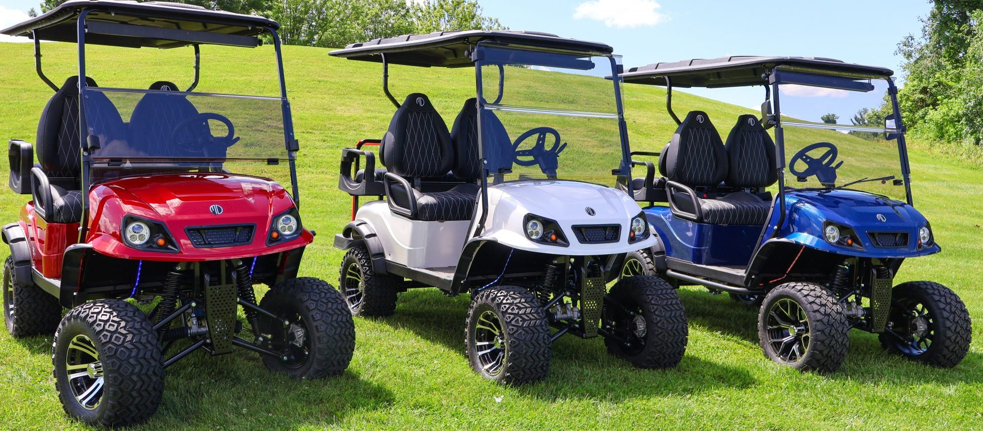 Three lifted golf carts—red, white, and blue—are parked side by side on green grass, with a grassy hill and trees in the background under a blue sky.