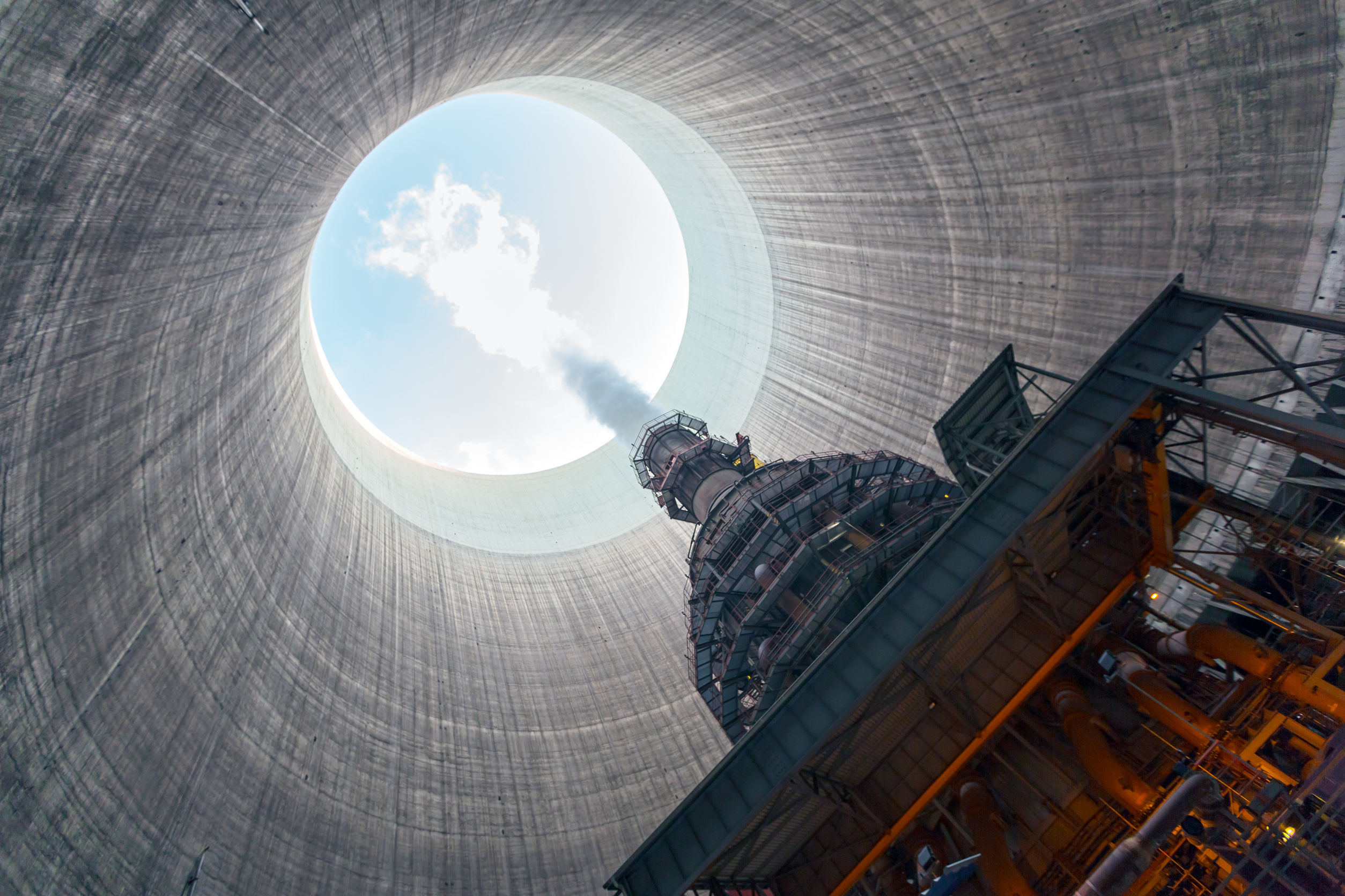 View from inside a large cooling tower, looking up towards the sky with clouds above. Steam rises from industrial equipment at the base, emphasizing the scale and height of the cylindrical concrete structure.