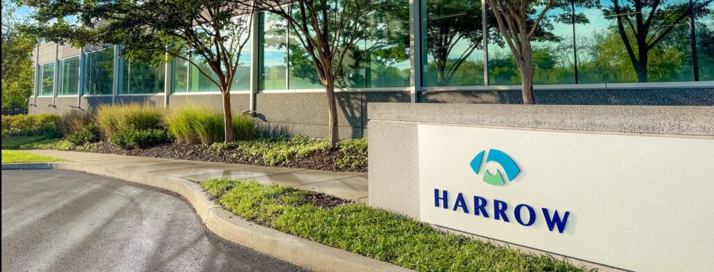 A modern office building with large windows and landscaped greenery. In the foreground, a white sign displays the blue and green Harrow logo and the word "HARROW" in blue letters.