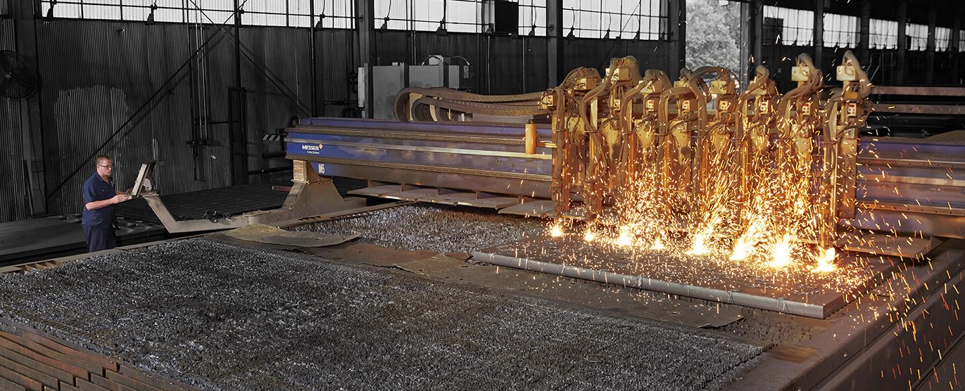 A worker operates a large industrial machine with multiple torches cutting metal sheets, producing bright sparks in a factory setting.