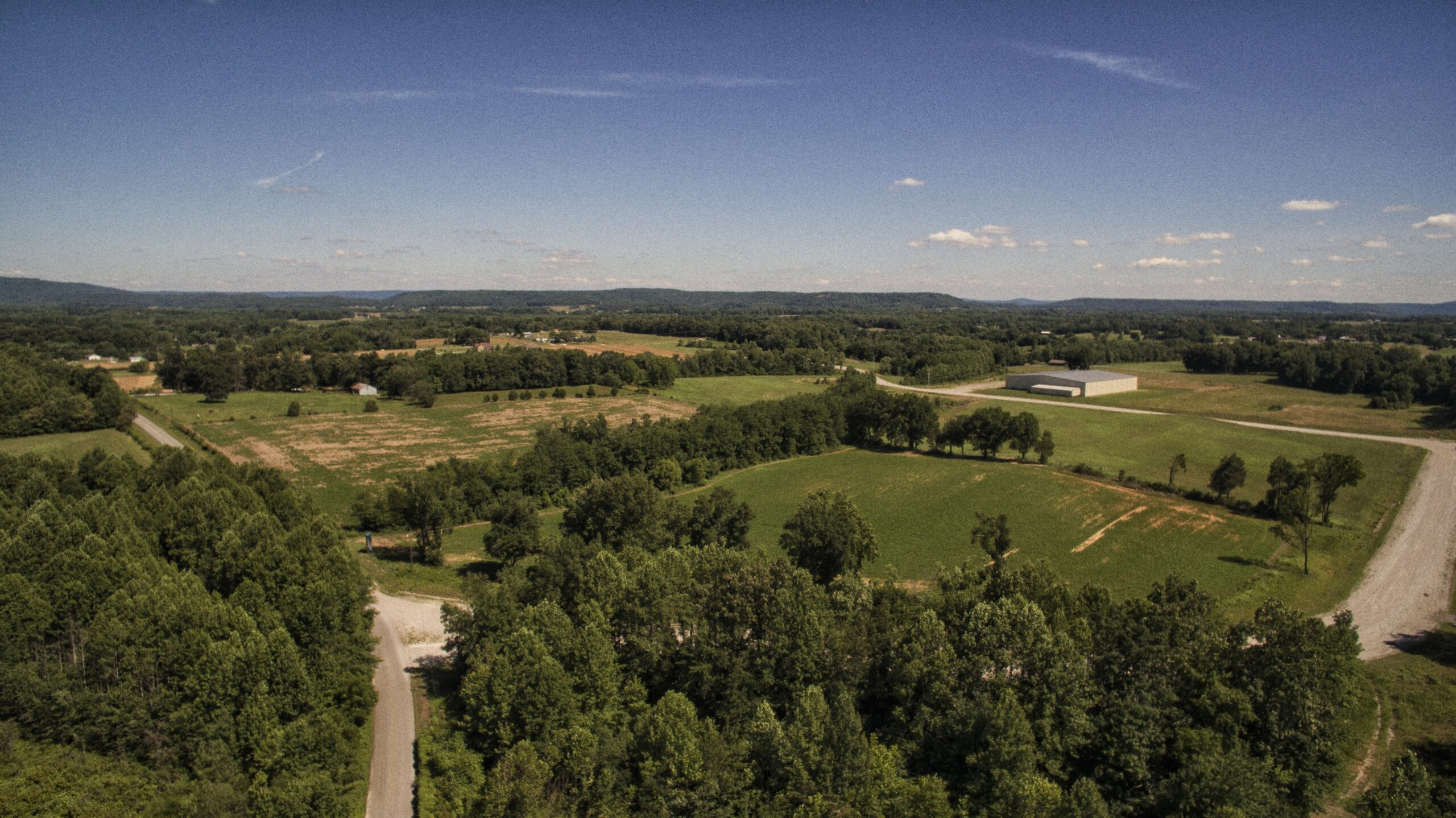 Aerial view of a rural landscape with green fields, scattered trees, a few roads, and a large building under a clear blue sky with distant hills in the background.
