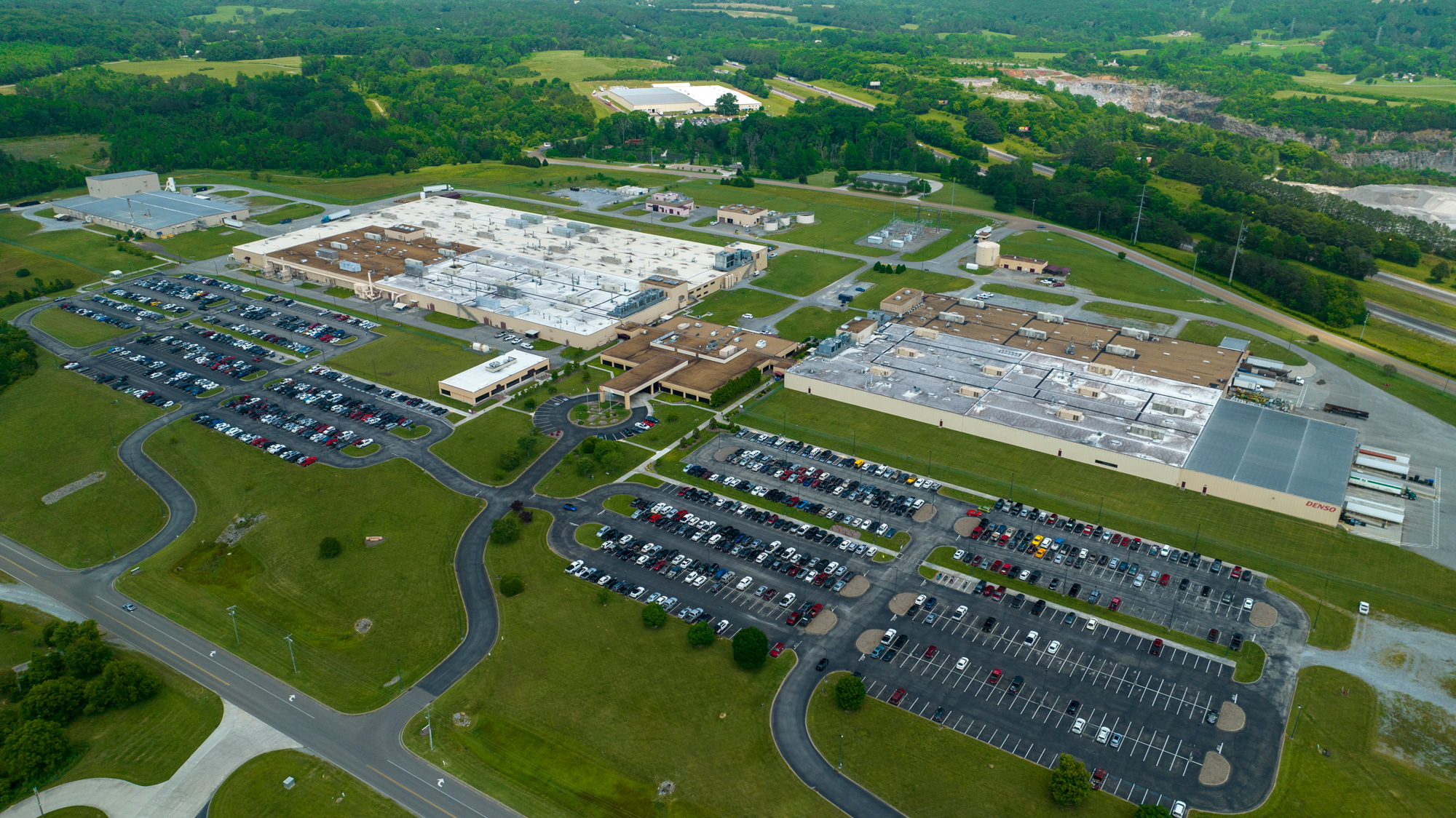 Aerial view of a large industrial facility with several connected buildings, multiple parking lots filled with cars, and green grassy areas surrounding the complex. Trees and roads are visible in the background.