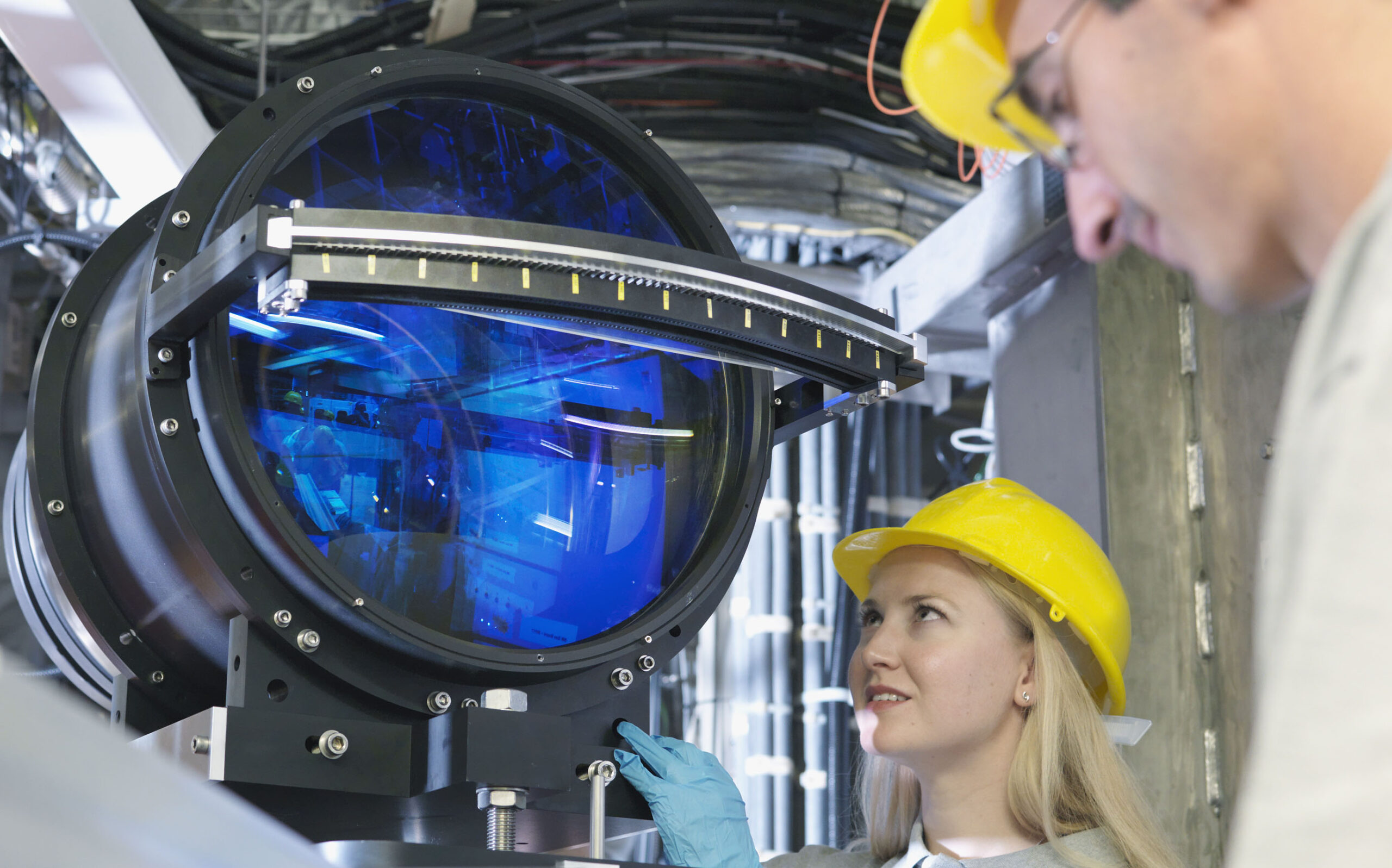Two engineers wearing yellow hard hats inspect a large, circular scientific instrument with a reflective blue surface, surrounded by cables and industrial equipment, in a high-tech lab setting.