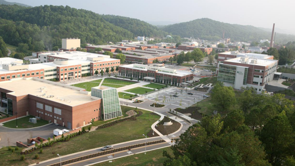 Aerial view of a sprawling campus with modern brick and glass buildings, pathways, green lawns, and surrounding wooded hills under a hazy sky. Roads and landscaped areas are visible in the foreground.