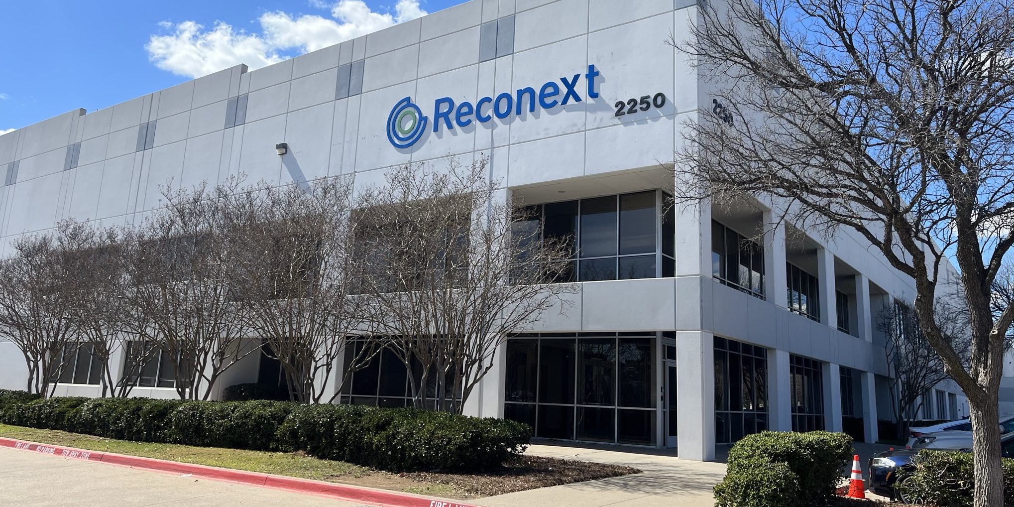 A modern two-story office building with the sign "Reconext" and street number 2250 on the facade, surrounded by leafless trees, shrubs, and a clear blue sky in the background.