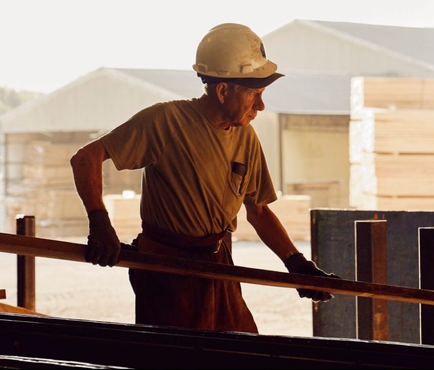 A man wearing a hard hat, gloves, and a T-shirt works outdoors, lifting a wooden plank at a lumber yard with stacks of wood and industrial buildings in the background.