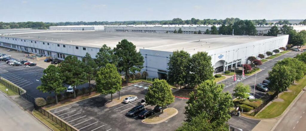 A large white warehouse building with rows of windows, surrounded by trees and parking lots with cars; the area appears clean and well-maintained under a clear sky.
