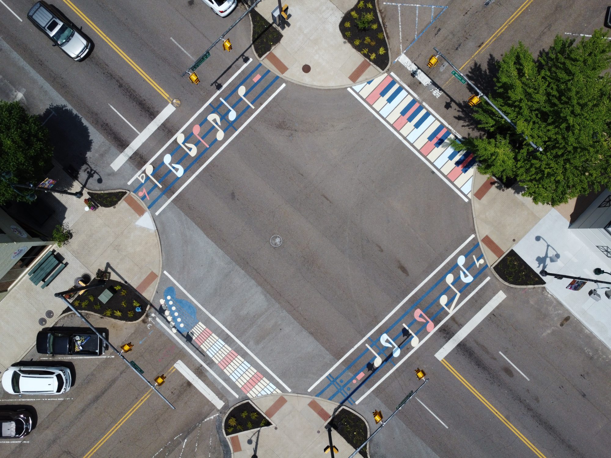 Aerial view of an intersection with crosswalks painted like piano keys, musical notes, and stars in red, white, and blue. Cars are parked along the street, and there are trees and sidewalks at the corners.