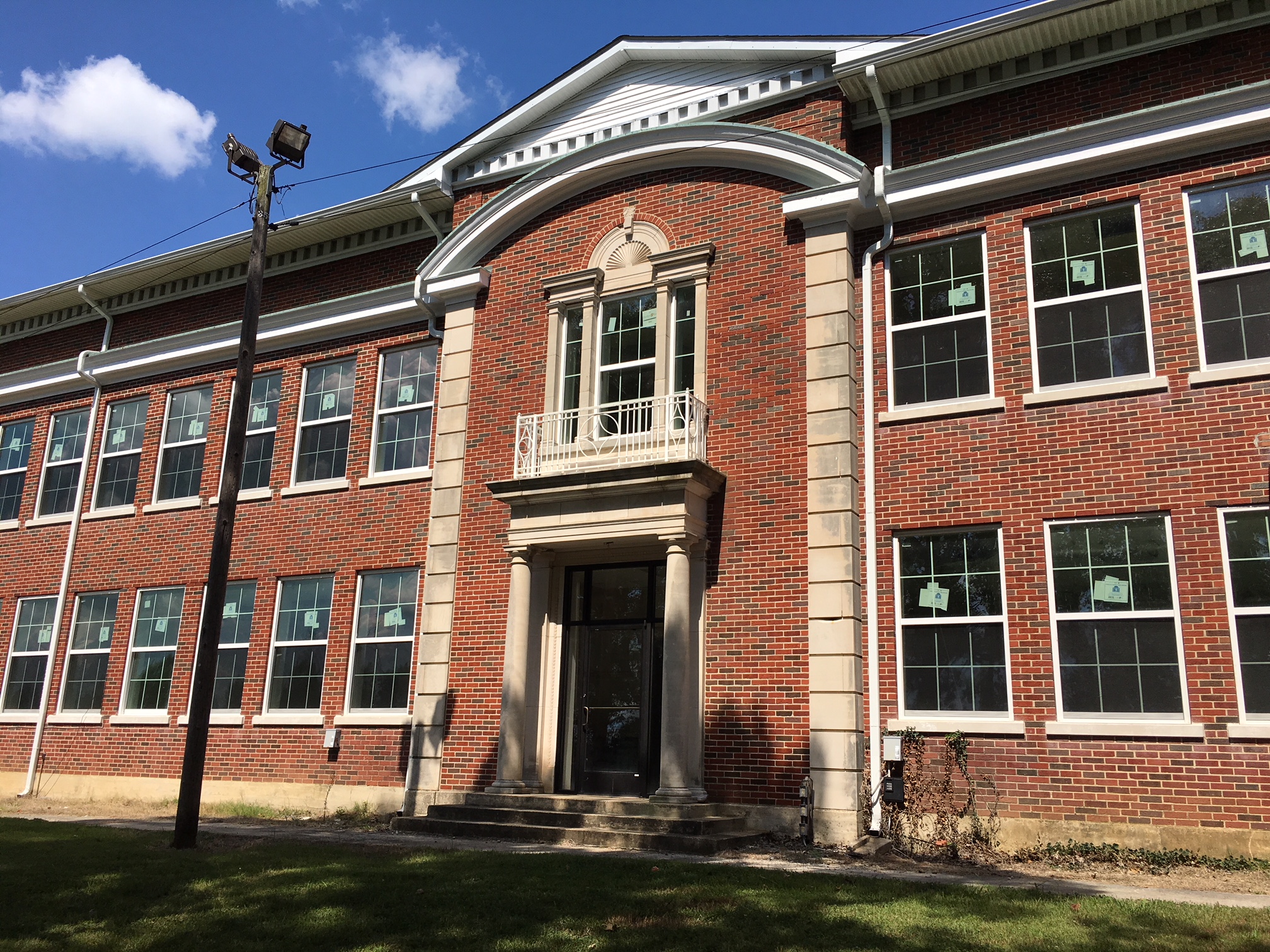 A red brick building with large windows, tall columns, and a decorative balcony above the main entrance, standing under a blue sky with scattered clouds.
