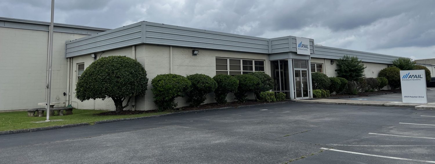 A low, light-colored commercial building with large windows, trimmed bushes along the front, and a sign reading “MAIL” near the entrance. The parking lot in front is mostly empty, and the sky is overcast.