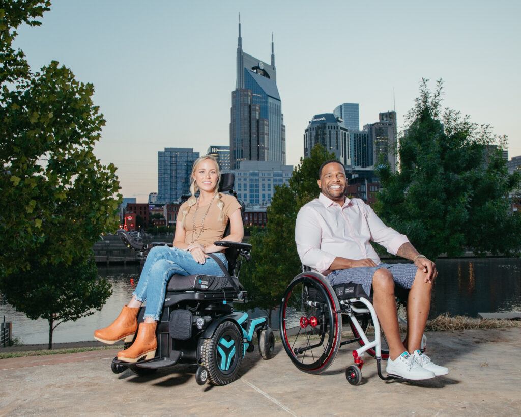 Two people in wheelchairs smiling outdoors near a river, with a city skyline and tall buildings in the background at dusk. Trees and water are also visible.