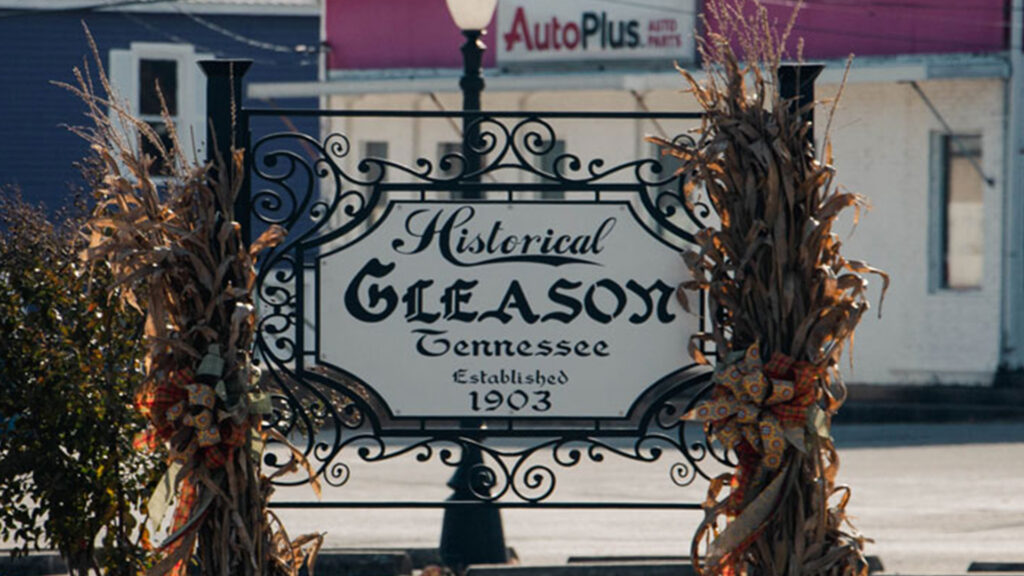 Decorative sign reading “Historical Gleason Tennessee, Established 1903,” framed by dried corn stalks and ribbons, with a street and Auto Plus storefront in the background.
