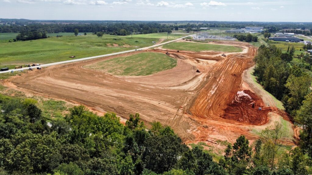 Aerial view of a large construction site with cleared earth, machinery tracks, and exposed red soil, surrounded by green fields, trees, and buildings in the distance under a partly cloudy sky.