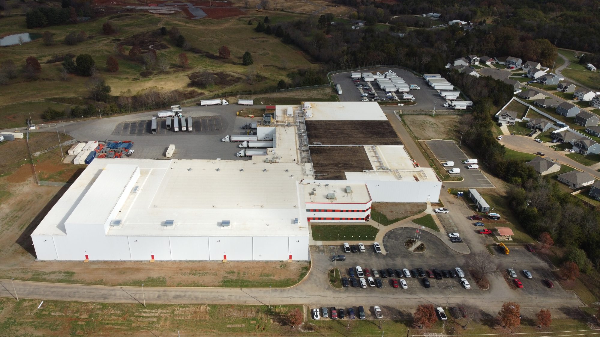 Aerial view of a large white industrial building with multiple loading docks, semi-trucks, parked cars, and nearby residential homes surrounded by trees and fields.