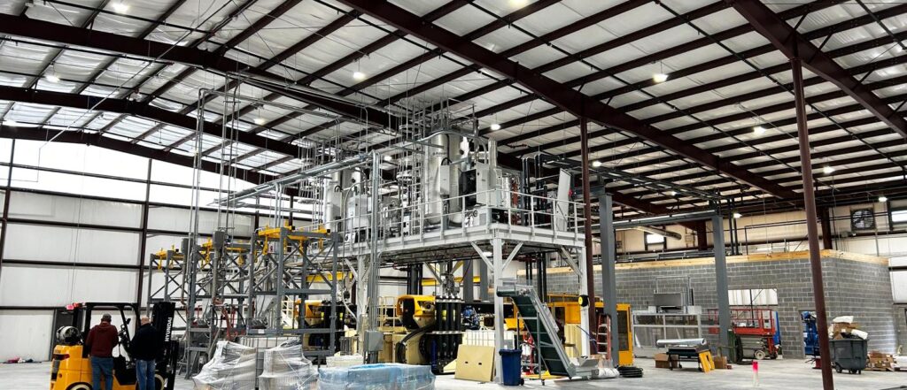 A spacious industrial warehouse with high ceilings, metal beams, and large machinery in the center, surrounded by equipment, pallets, and construction materials. Workers and a forklift are visible near the machinery.