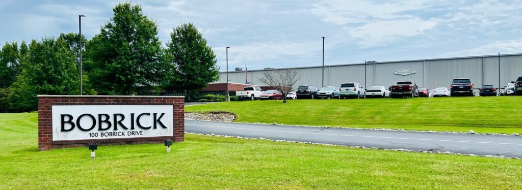 A grassy lawn with a brick sign reading "Bobrick, 100 Bobrick Drive" in front of a large industrial building with parked cars and trees under a cloudy sky.