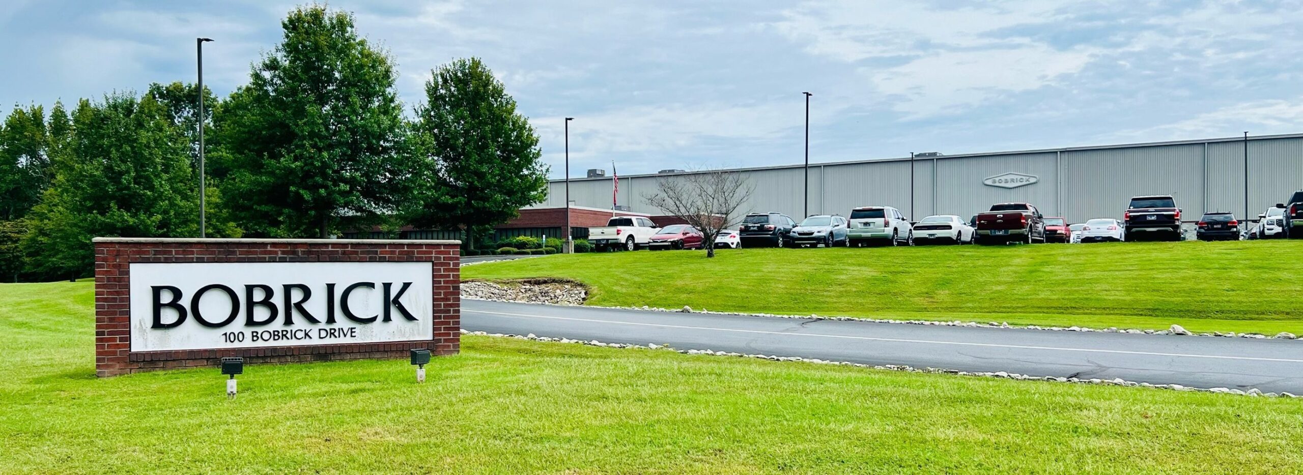 A grassy lawn with a brick sign reading "Bobrick, 100 Bobrick Drive" in front of a large industrial building with parked cars and trees under a cloudy sky.