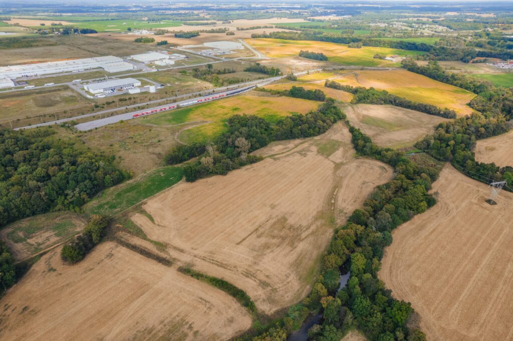 Aerial view of a rural landscape with harvested fields, patches of trees, a small stream, a road with trucks, and industrial buildings in the background under a partly cloudy sky.