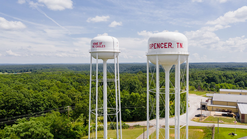 Aerial view of two white water towers labeled "SPENCER, TN" and "HOME OF THE EAGLES," standing above a forested landscape and buildings under a partly cloudy sky.