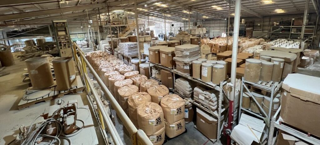 A large warehouse filled with stacks of brown paper bags, cardboard barrels, and boxes containing powdery materials, arranged neatly on metal shelves under a high ceiling with bright lights.