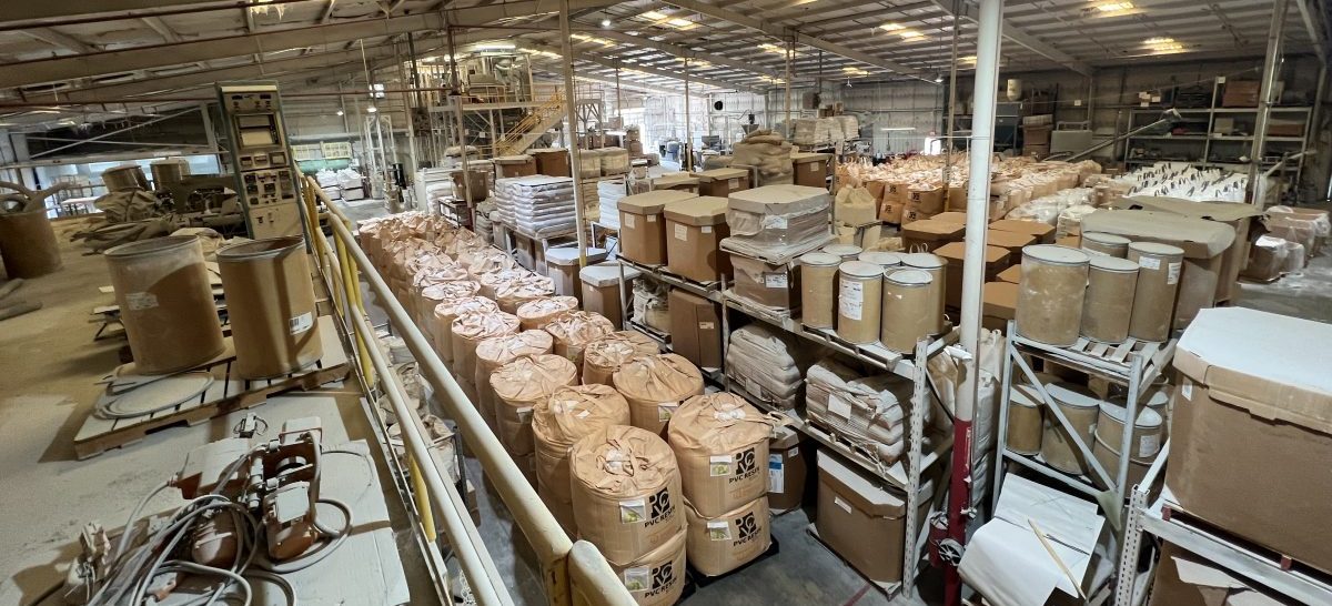 A large warehouse filled with stacks of brown paper bags, cardboard barrels, and boxes containing powdery materials, arranged neatly on metal shelves under a high ceiling with bright lights.