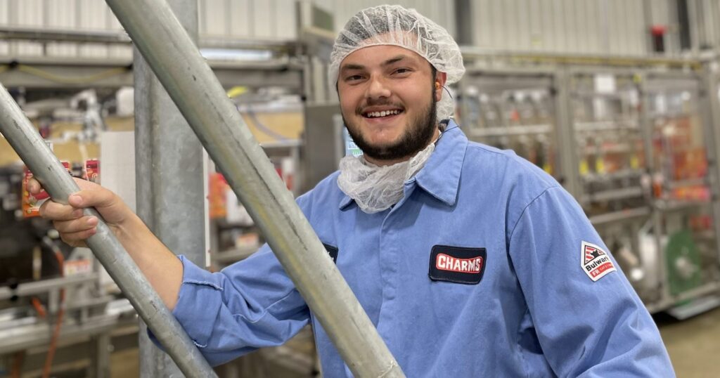 A smiling person wearing a blue work uniform, hairnet, and beard cover stands in a factory or industrial setting, holding onto a metal railing. Machinery and equipment are visible in the background.
