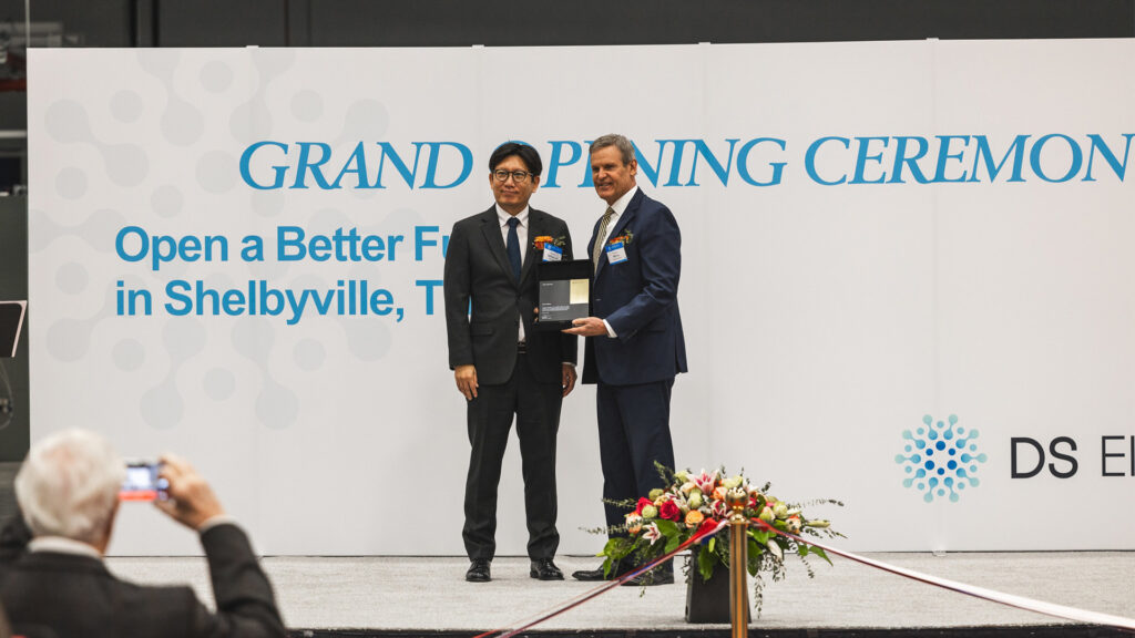 Two men in suits stand on stage during a grand opening ceremony. One man holds a plaque. A flower arrangement and red ribbon are at their feet. The background displays event text and the DS E logo. An audience member takes a photo.