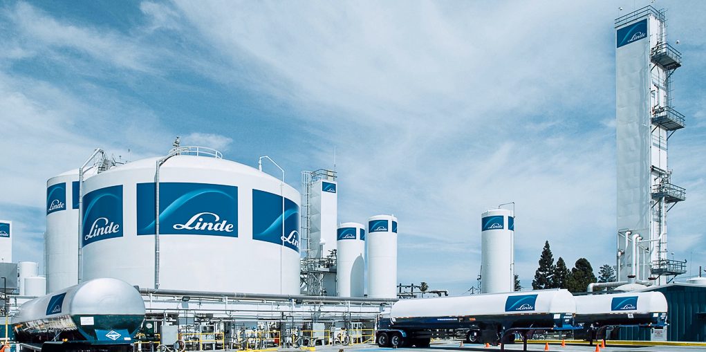 Industrial facility with multiple large white tanks, pipes, and tall structures displaying the "Linde" logo, set under a blue sky with scattered clouds. A white tanker truck is parked in the foreground.