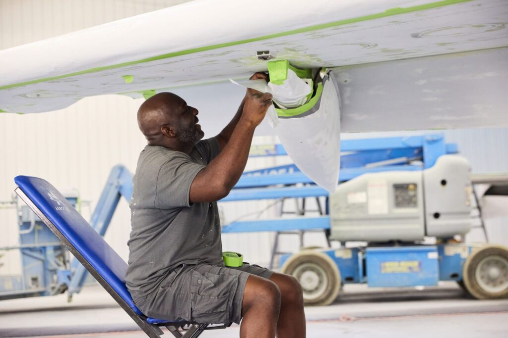 A man sits on a blue chair, taping and prepping the underside of an aircraft wing for painting inside a hangar, with equipment visible in the background.
