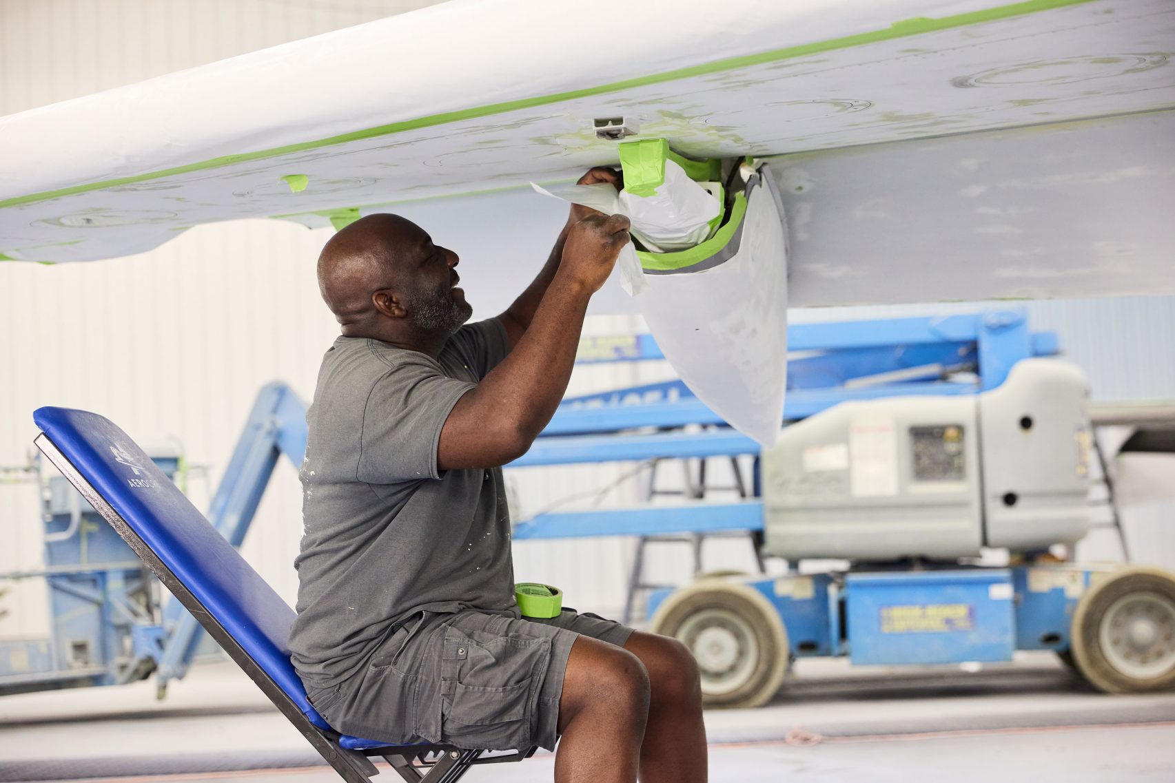 A man sits on a blue chair, taping and prepping the underside of an aircraft wing for painting inside a hangar, with equipment visible in the background.