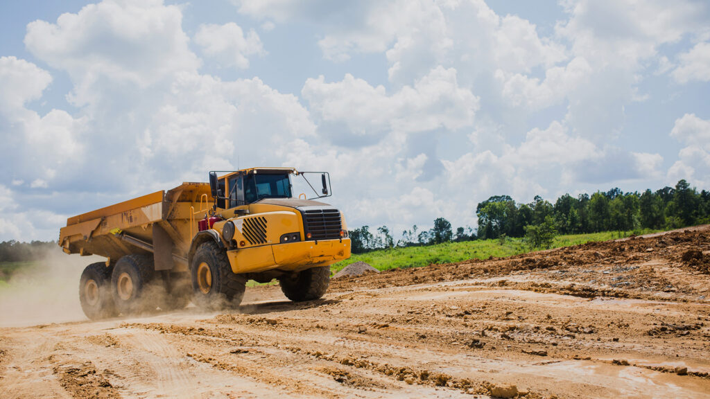 A large yellow dump truck drives on a dusty construction site under a partly cloudy sky, with trees and greenery visible in the background.