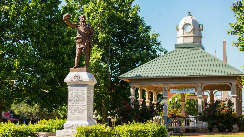 A bronze statue of a man raising his hat stands on a stone pedestal in a park, near a gazebo with a green roof and a clock tower. Lush trees and blooming bushes surround the area. A sign reads “Lawrenceburg, TN.”.