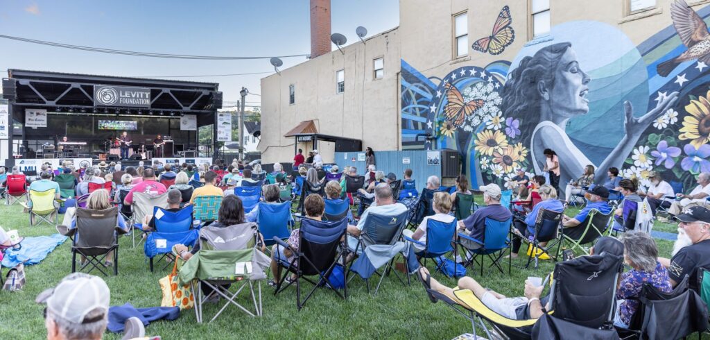 A crowd sits on lawn chairs facing an outdoor stage with live music. A large mural of a woman with butterflies and flowers decorates the wall beside the audience. The event takes place on a grassy area near buildings.